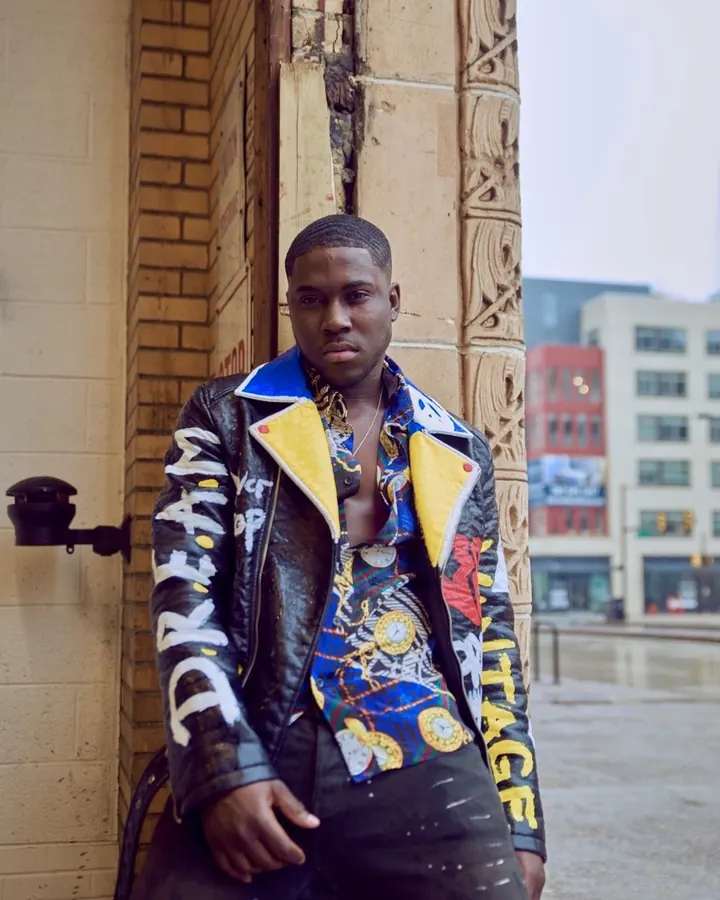 Homme torse nu, peau foncée, portant une veste en cuir noire avec col jaune, chemise colorée ouverte, regardant vers le bas, devant un mur en briques.