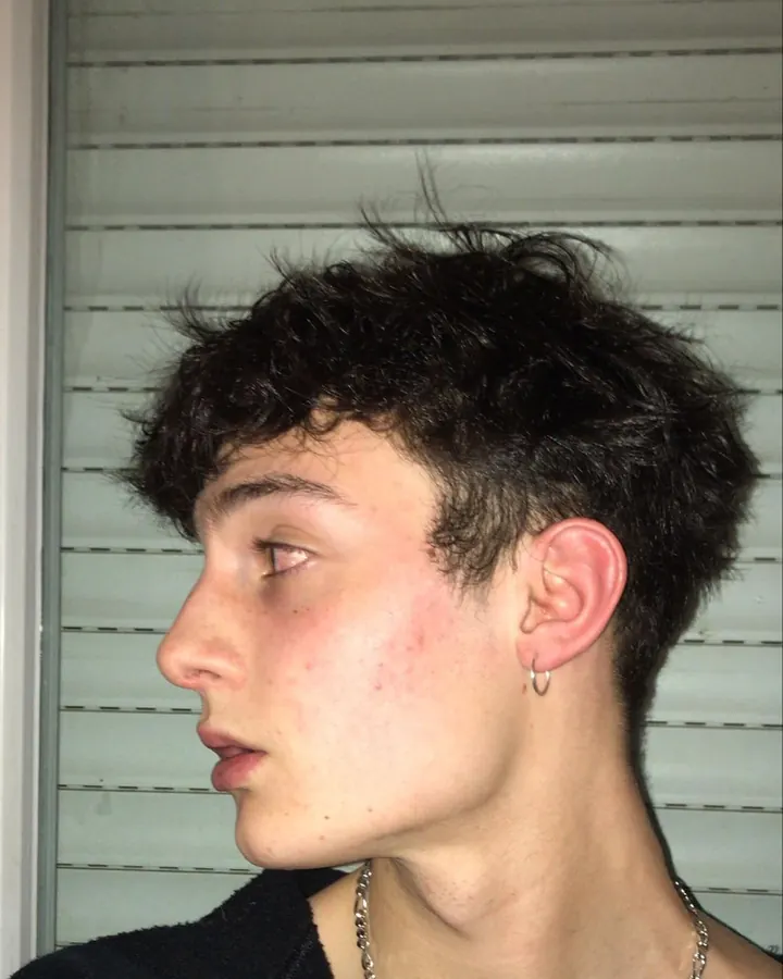 Profil d’un jeune homme aux cheveux noirs bouclés, oreille percée, regard vers le côté, devant des volets blancs.