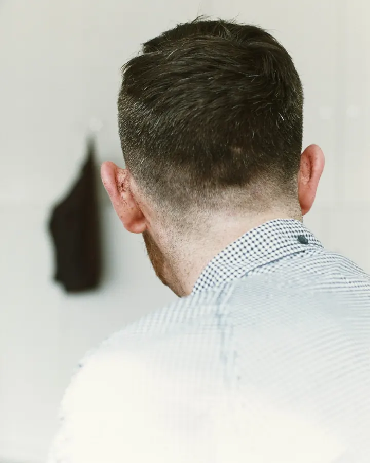 Vue arrière d’un homme aux cheveux courts, portant une chemise à carreaux blancs et bleus, regardant vers le côté, dans une pièce claire.