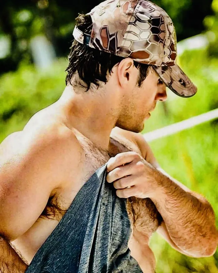 Homme torse nu, barbe courte, portant une casquette camouflage, tenant un tissu sur son épaule, regard vers le côté, en plein air verdoyant.
