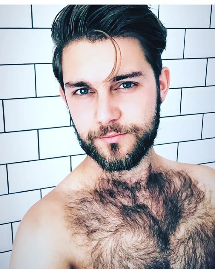 Homme barbu, torse nu, portant un débardeur noir, faisant un geste de poing devant sa bouche, souriant, dans une salle de sport.