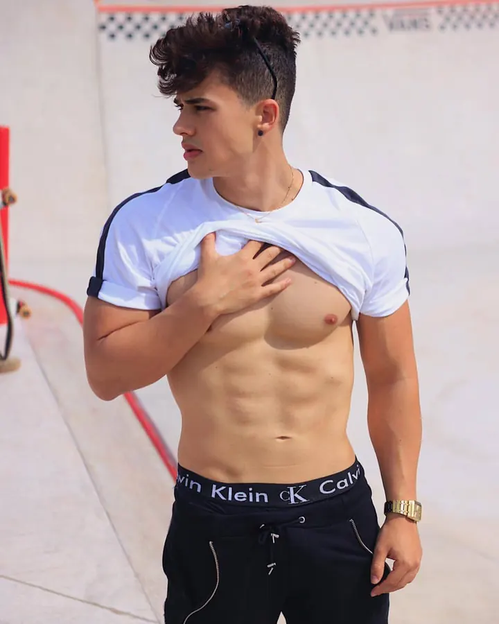 Jeune homme torse nu, portant un short blanc, assis sur un lit rouge, tenant un téléphone avec une coque colorée, main derrière la tête, regard vers le bas.