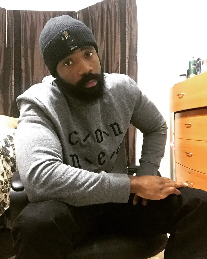 Homme torse nu, peau foncée, portant une casquette bleue, tenant un appareil photo vintage devant lui, en sous-vêtements rayés gris, devant un rideau beige.