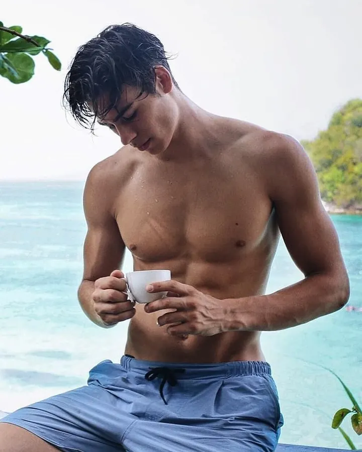 Jeune homme torse nu, portant un short blanc, tenant une paire de baskets blanches par les lacets, buvant de l’eau, devant un mur blanc.