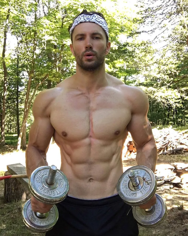 Homme européen torse nu, musclé, en forêt, tenant deux haltères, bandeau sur la tête, regard direct vers l'appareil.