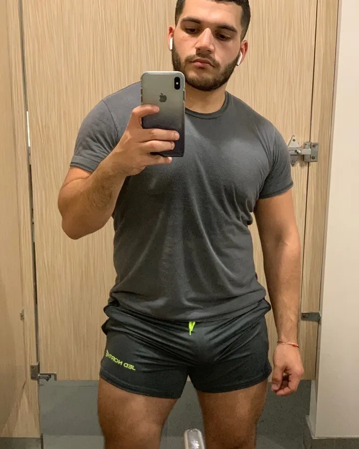Homme européen en t-shirt gris, barbe courte, prenant un selfie dans un miroir, short noir, écouteurs blancs, regard concentré.