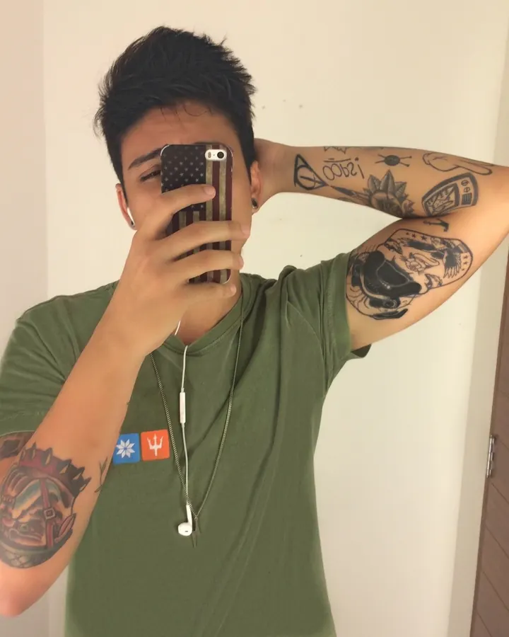 Jeune homme européen en t-shirt vert, tatouages visibles aux bras, prenant un selfie dans un miroir, écouteurs blancs, téléphone avec coque drapeau américain.