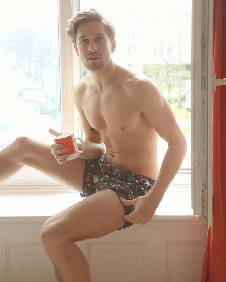 Homme européen torse nu, assis sur un rebord de fenêtre, tasse rouge à la main, boxer coloré, regard direct, lumière naturelle.