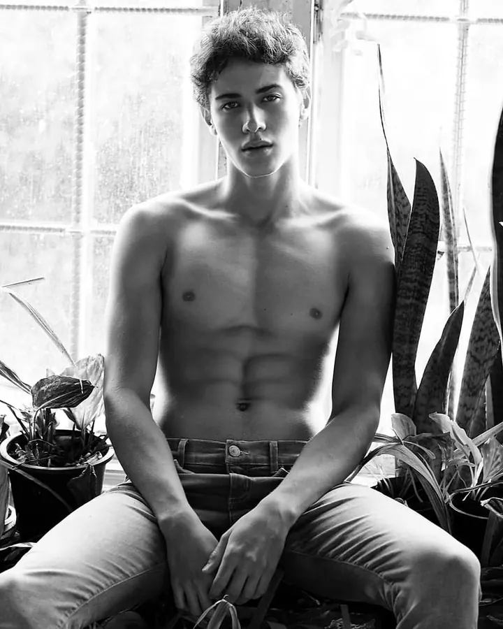 Jeune homme torse nu, peau blanche, cheveux bouclés, assis parmi des plantes, regard direct, jeans clairs.