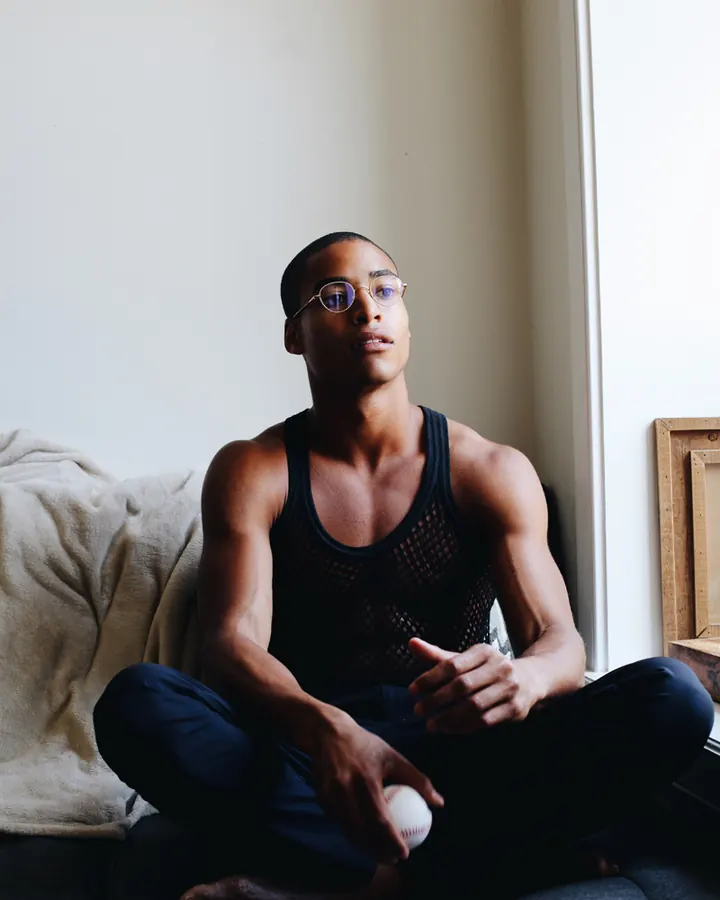 Homme torse nu, peau foncée, lunettes rondes, maille noire, assis en tailleur sur un canapé, tenant une balle de baseball.