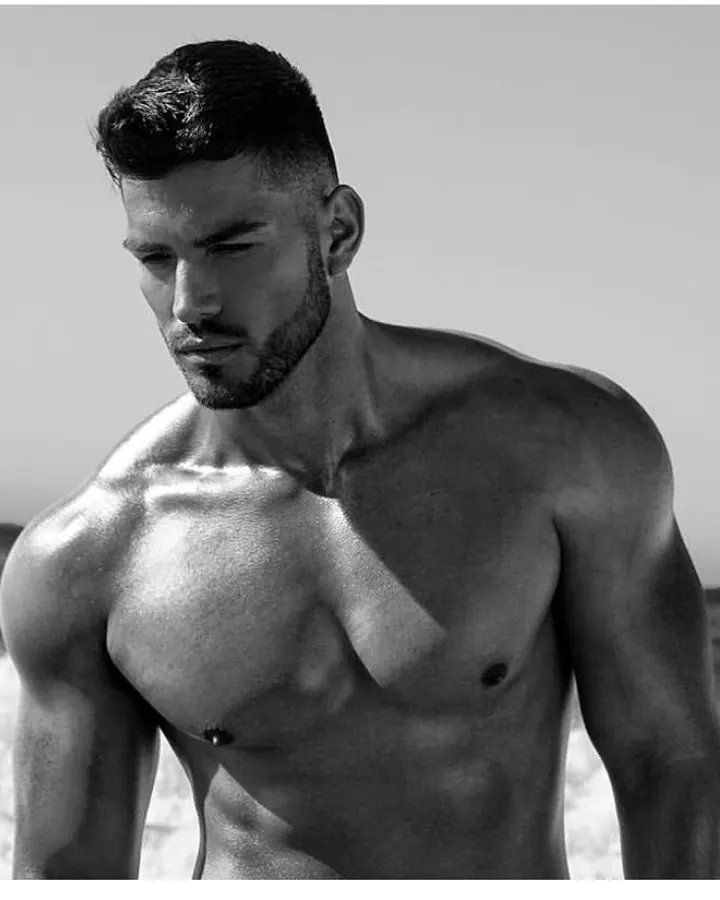 Homme torse nu, peau bronzée, barbe courte, cheveux courts, regard vers le bas, photo en noir et blanc, plage.