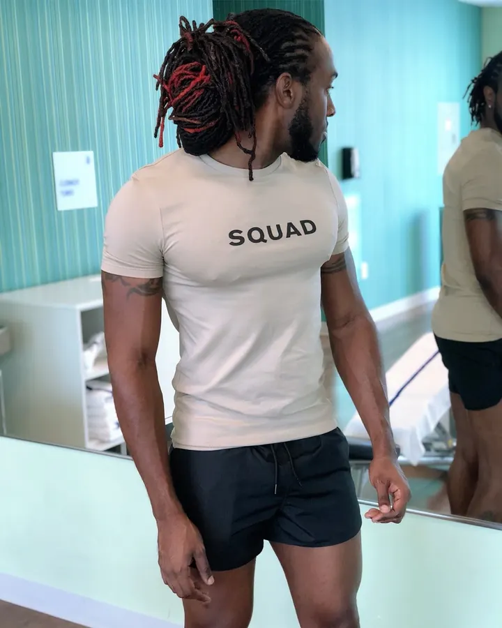 Homme torse nu, peau foncée, dreadlocks rouges et noires, t-shirt beige 'SQUAD', shorts noirs, regard de profil.