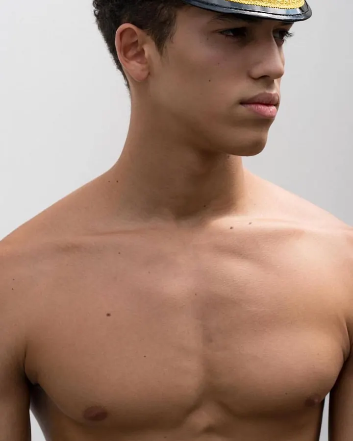 Jeune homme torse nu, peau blanche, cheveux bruns-noirs courts, portant une casquette de capitaine, regard de profil.