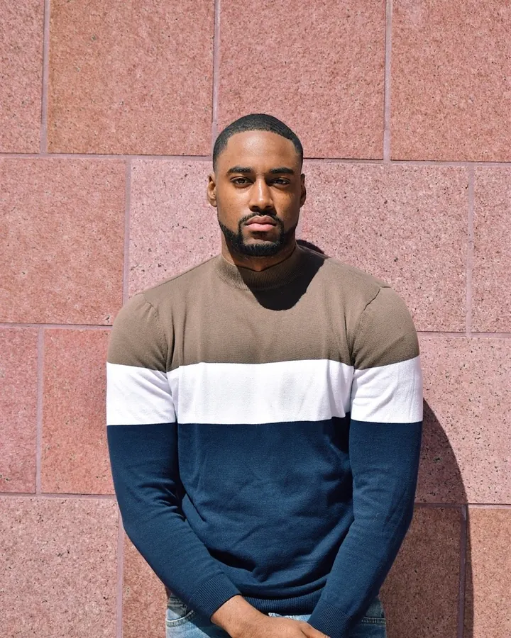 Homme torse nu, peau foncée, barbe courte, cheveux courts, regard direct, vêtu d'un pull tricolore, contre un mur rose.