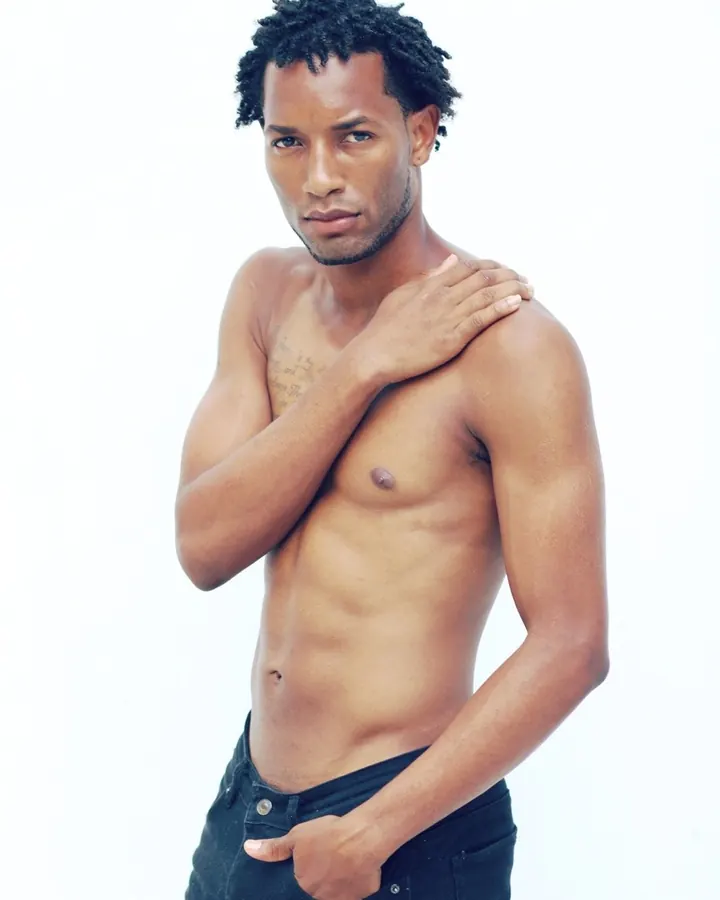 Homme torse nu, peau mate, cheveux crépus noirs, main sur l'épaule, regard direct, pantalon noir.