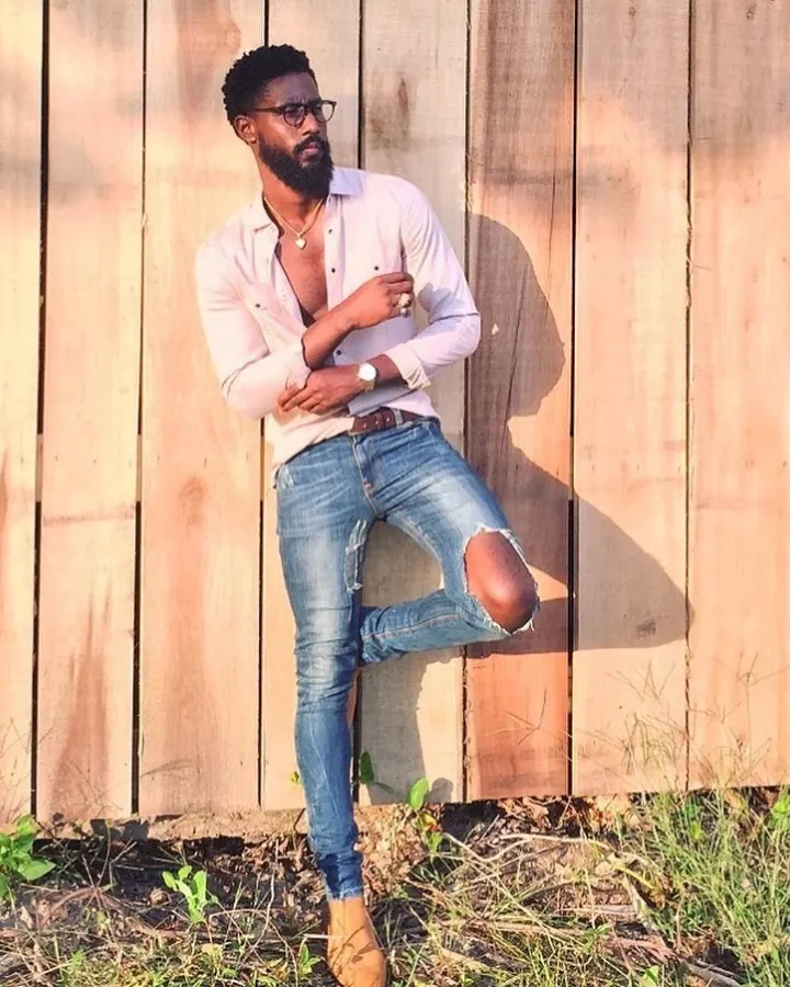Homme en chemise rose clair boutonnée, jeans déchirés, lunettes, chaussures marron, appuyé contre un mur en bois.