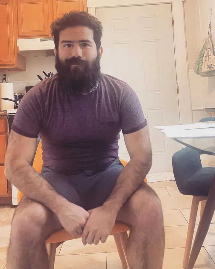 Homme assis à une table de cuisine, t-shirt violet, barbe fournie, regardant vers le haut.