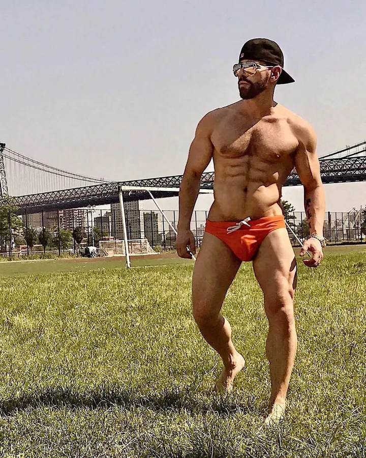 Homme torse nu, barbe courte, regard intense, en plein air, avec un pont en arrière-plan, sous-vêtement orange.