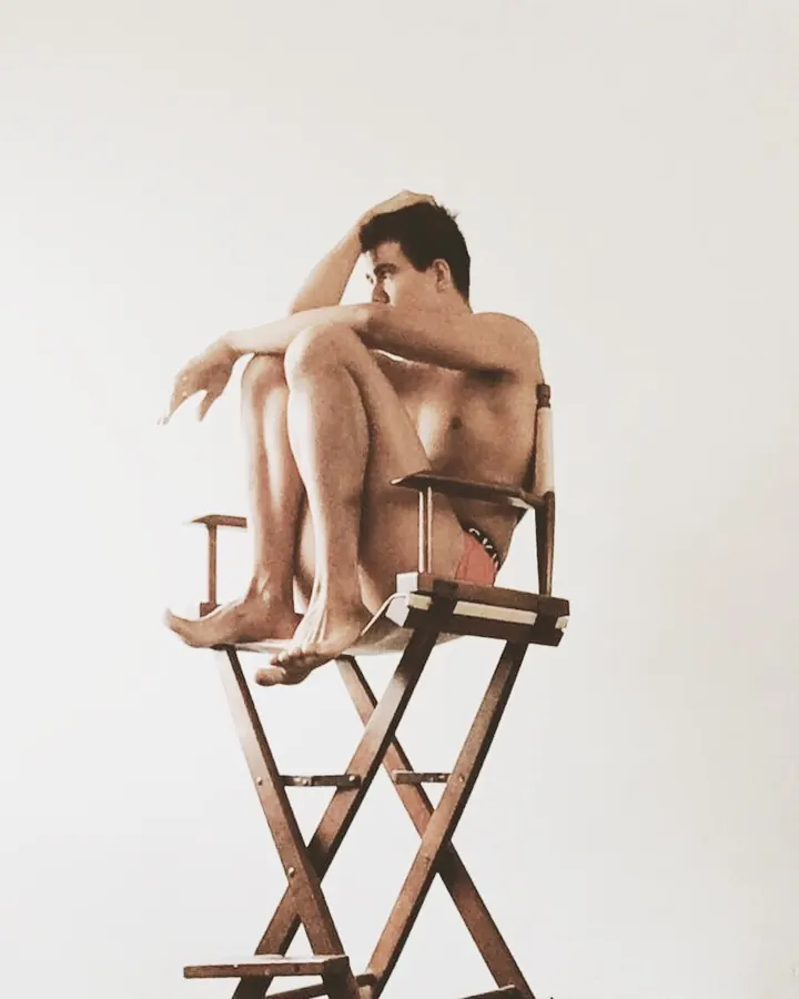 Homme torse nu, assis sur une chaise haute en bois, les jambes croisées, regardant de côté.
