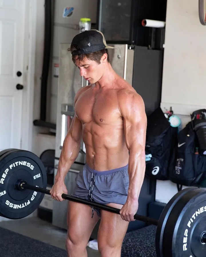 Homme torse nu, barbe courte, regard intense, en salle de sport, soulevant une barre de poids.