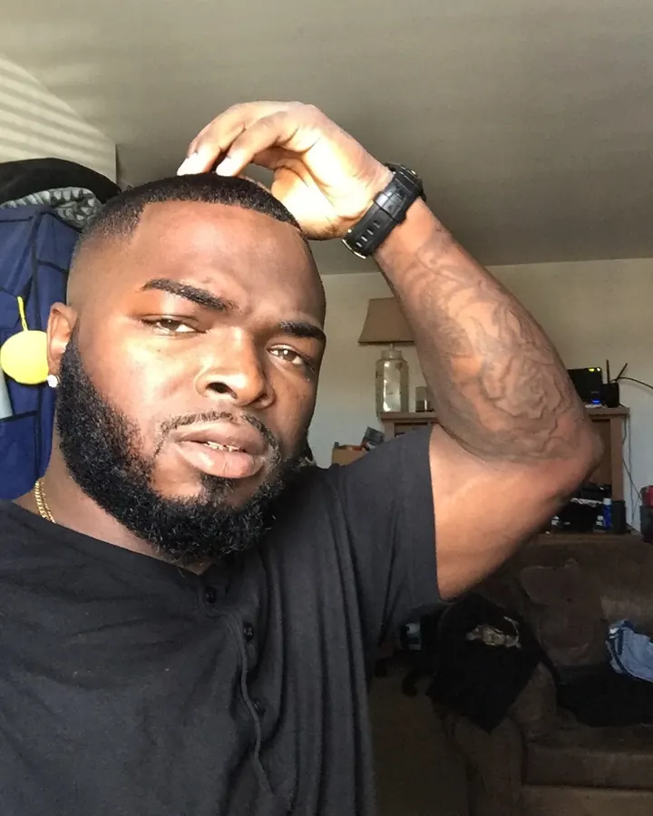 Homme en t-shirt noir, barbe fournie, regard intense, dans une pièce intérieure, main sur la tête.
