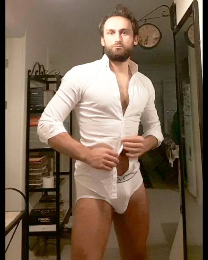Homme en chemise blanche boutonnée, sous-vêtement blanc, tenant le bas de sa chemise, dans une pièce intérieure.