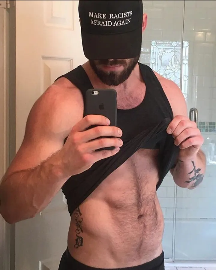 Homme en tank top noir, capuche noire avec inscription 'MAKE RACISTS AFRAID AGAIN', prenant un selfie dans un miroir de salle de bain.