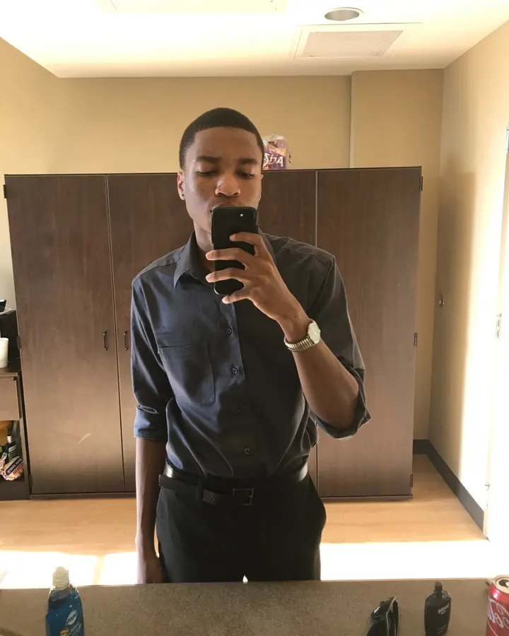 Homme en chemise sombre et pantalon noir, prenant un selfie dans un miroir d'une salle de bain.