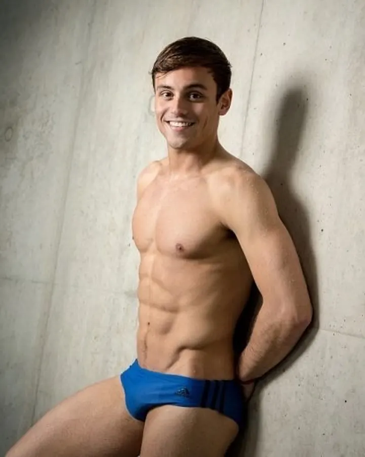 Homme torse nu, en sous-vêtements bleus, appuyé contre un mur de béton, souriant vers l’appareil photo.