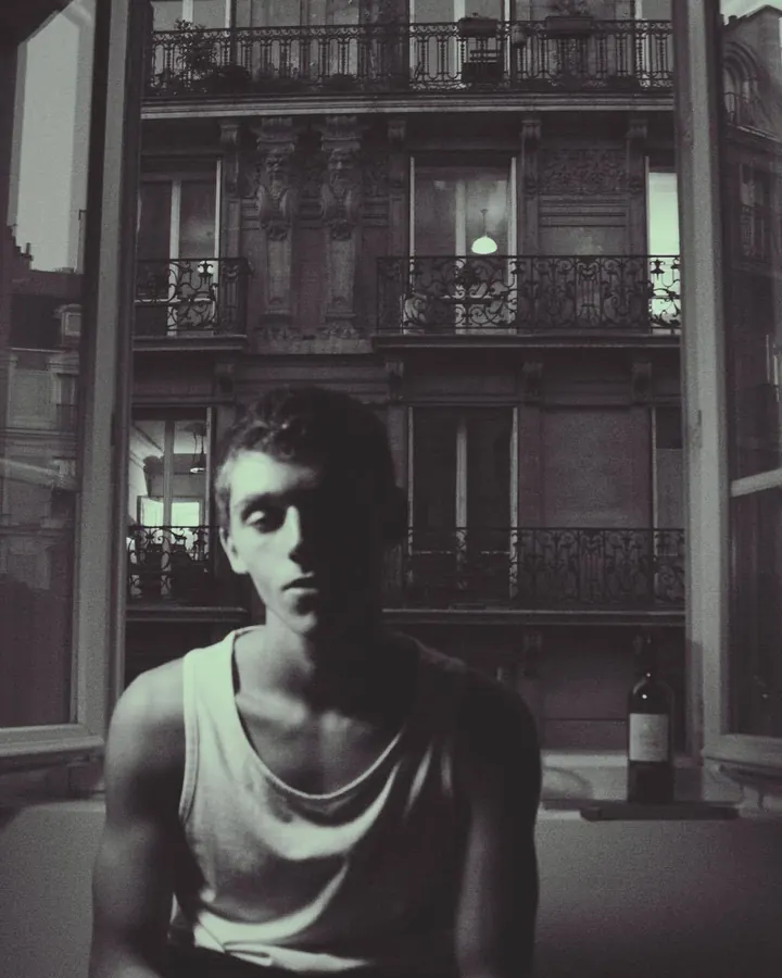 Homme en sous-vêtement blanc, debout près d'une fenêtre, regardant vers l'appareil photo, dans une ambiance naturelle et douce.