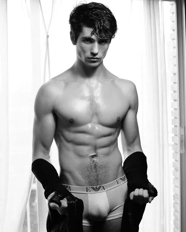 Homme torse nu, en sous-vêtements, tenant un vêtement noir, debout devant un rideau blanc, regard intense.