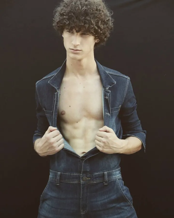 Homme torse nu, en jean, ouvrant sa veste en denim, regardant vers l’appareil photo, sur fond noir.