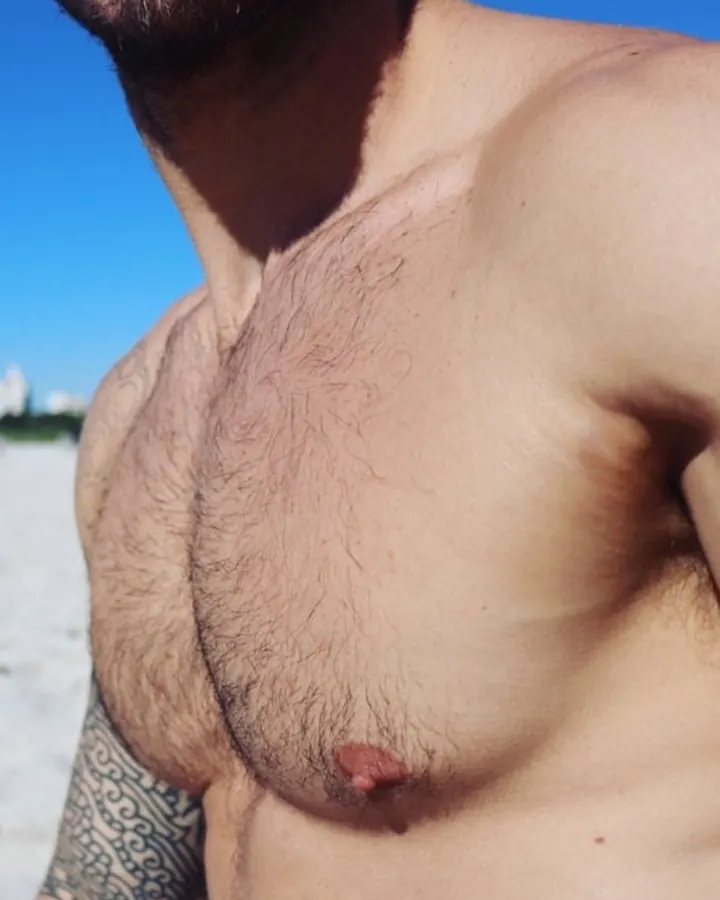 Homme torse nu sur la plage, barbe, tatouage au bras, ciel bleu en arrière-plan
