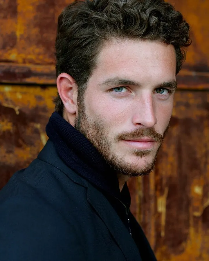 Homme aux yeux bleus, barbe courte, vêtu d’un manteau noir, regard intense contre un fond rouillé.