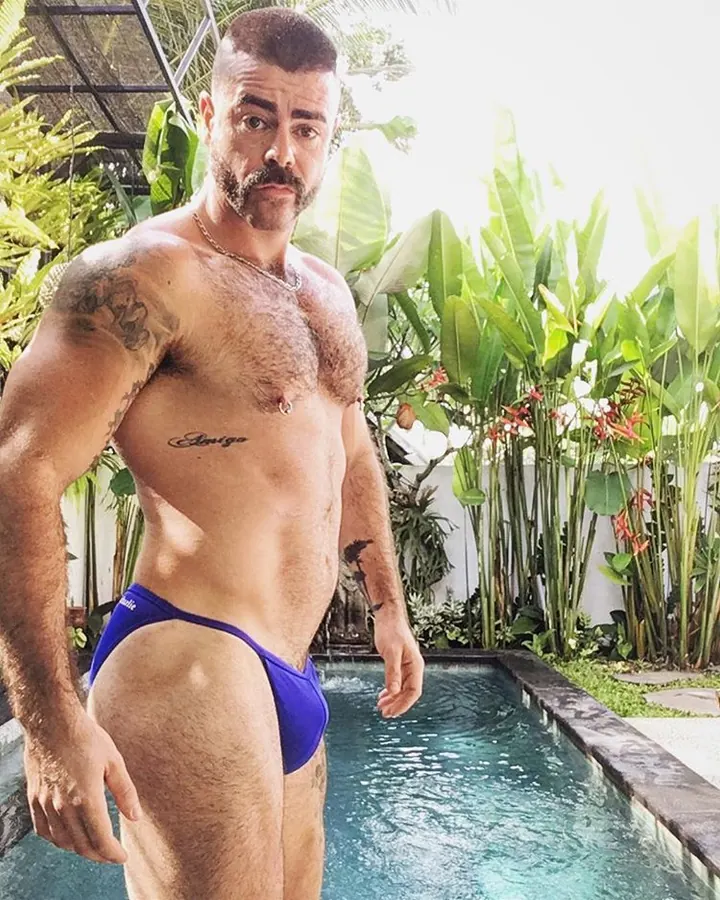 Homme barbu, regardant vers le côté, portant une chemise blanche légèrement ouverte, devant une fenêtre.