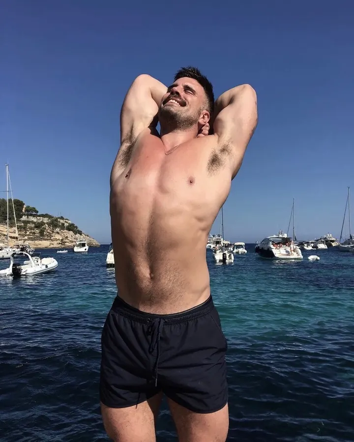 Homme barbu, torse nu, portant un short noir, les mains derrière la tête, souriant, debout devant une mer avec des bateaux.