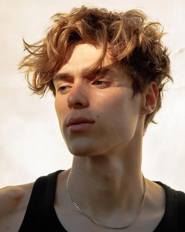 Jeune homme aux cheveux blonds, regardant vers le côté, avec un collier fin autour du cou, sur un fond lumineux.