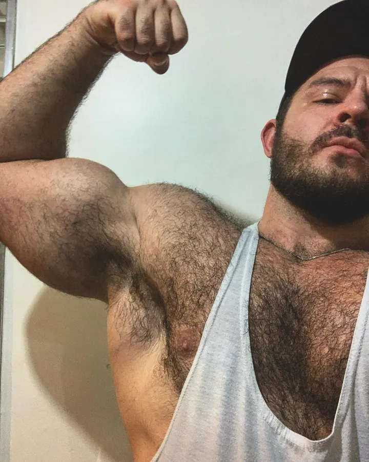 Homme barbu, torse nu, portant un débardeur blanc et un bonnet noir, montrant son biceps gauche en serrant le poing.
