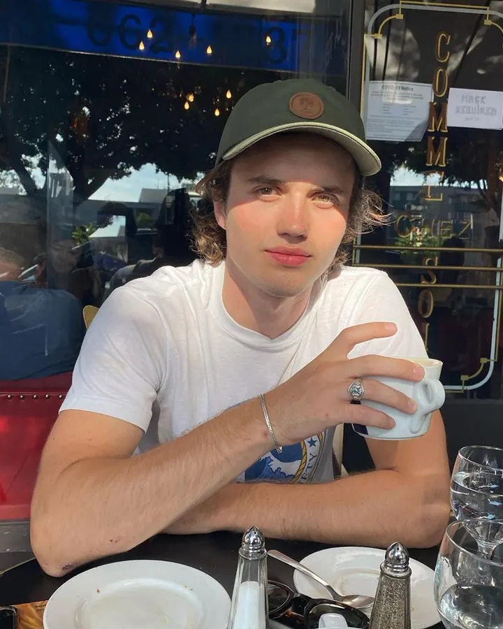 Jeune homme aux cheveux châtains portant un bonnet vert, assis à une table de café avec une tasse blanche en main.