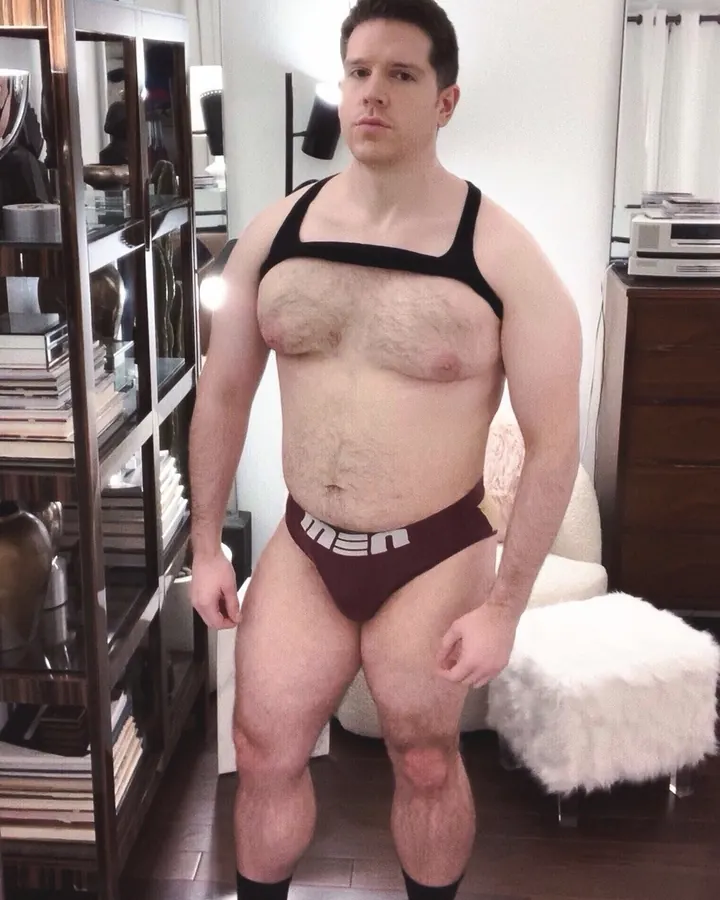 Homme barbu, regardant vers le côté, portant une chemise blanche légèrement ouverte, devant une fenêtre.