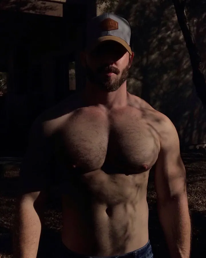 Homme européen aux cheveux châtains et barbe, torse nu, portant une casquette, dans une ambiance ombragée, en plan moyen.