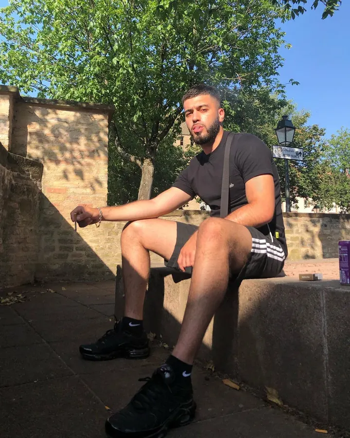 Homme européen aux cheveux châtains et barbe, assis sur un mur en plein air, vêtu d’un t-shirt noir et de shorts, sous un arbre verdoyant.