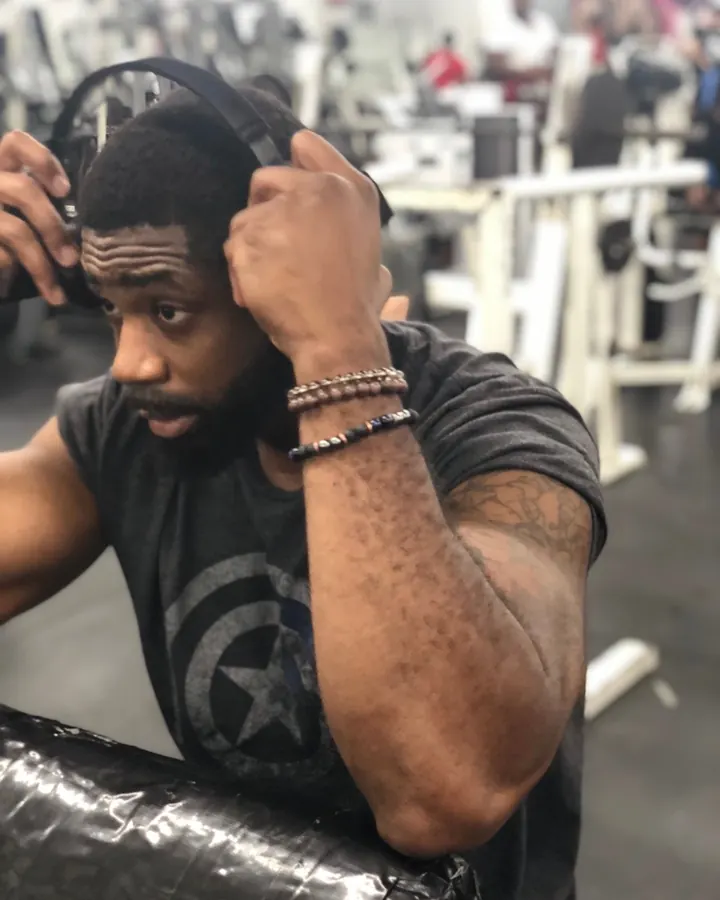 Homme africain de 35 à 45 ans, en salle de sport, portant un t-shirt noir, mettant ses écouteurs, tatouage sur le bras droit.