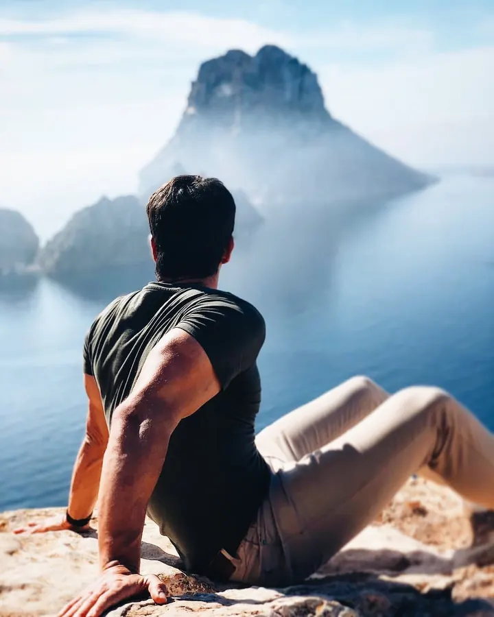Homme européen de 25 à 35 ans, vu de dos, assis sur un rocher, regardant une île lointaine, vêtu d'un t-shirt noir et de pantalons clairs.