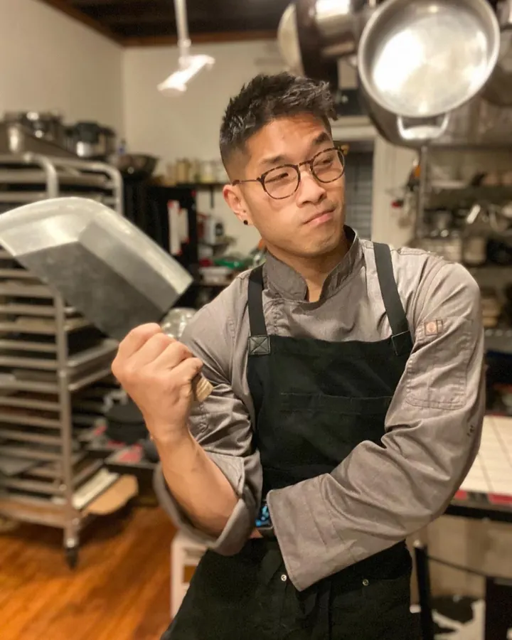 Homme asiatique en tenue de chef, lunettes, tenant une poêle dans une cuisine professionnelle.