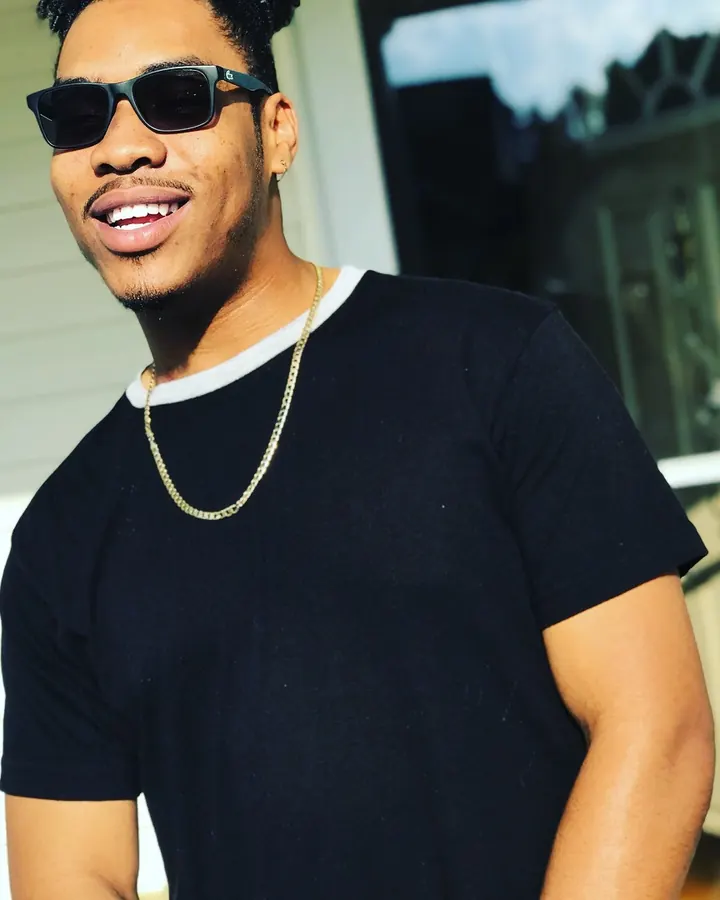 Homme africain souriant avec lunettes de soleil, t-shirt noir et chaîne dorée, en extérieur.