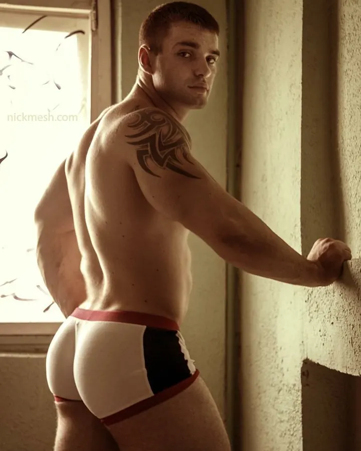 Homme européen torse nu, musclé, avec tatouage tribal sur l'épaule, en sous-vêtements blancs et rouges, adossé à un mur.