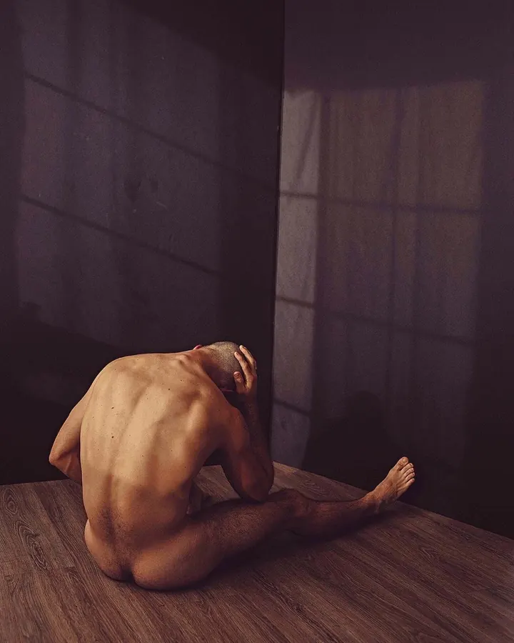Homme torse nu, assis sur le sol, la tête baissée, les mains sur la tête, dans un coin sombre.