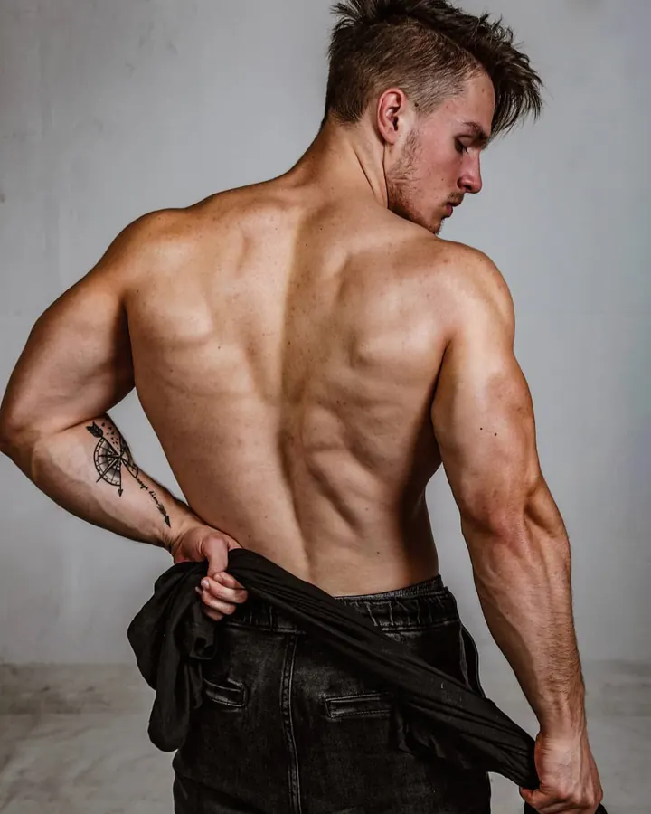 Homme torse nu, dos tourné, tenant un vêtement noir, tatouage visible sur le bras gauche, devant un mur gris.