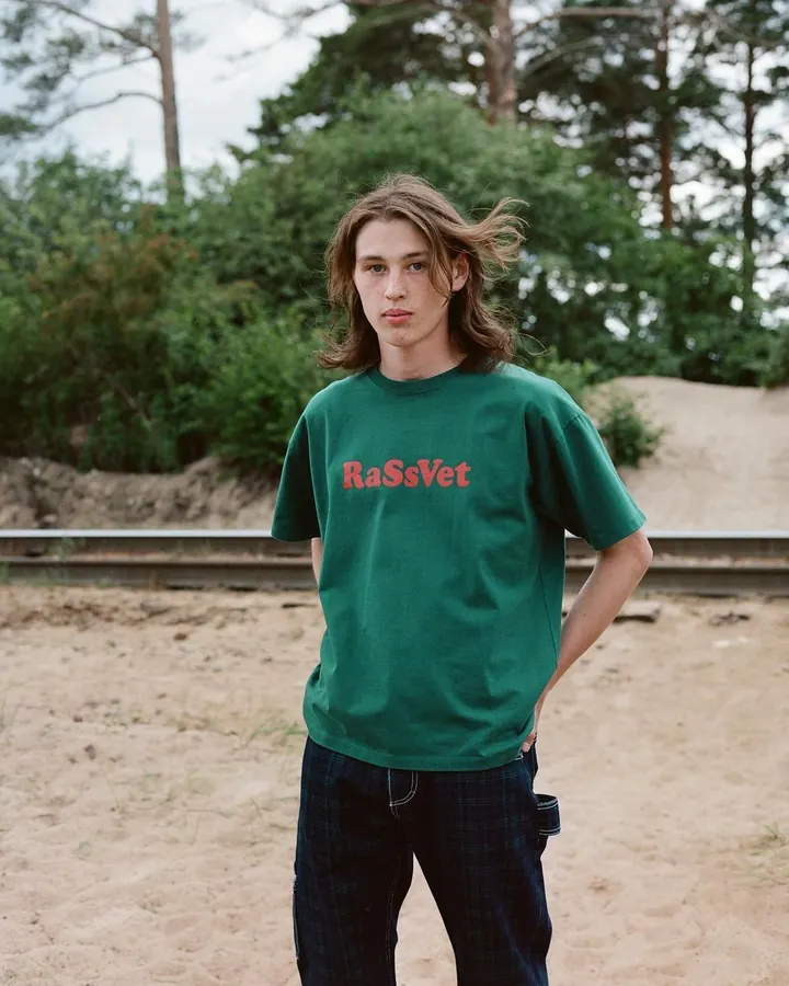 Jeune homme aux cheveux châtains portant un t-shirt vert avec l'inscription RaSsVet, debout dans un environnement extérieur avec des arbres en arrière-plan.